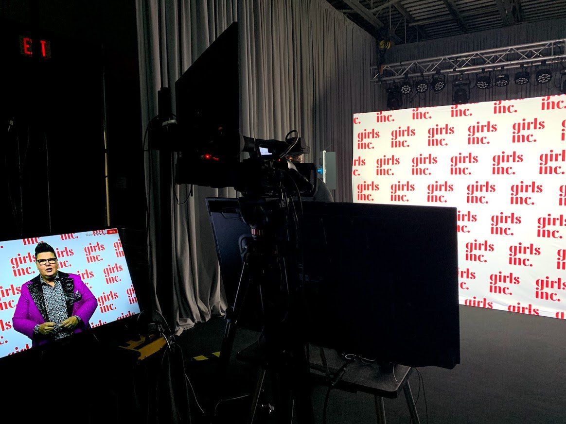 A person on a monitor is in front of a “Girls Inc.” backdrop. There are cameras and monitors set up in a studio.