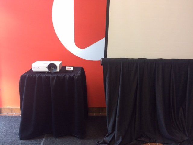 White projector on a black draped table against a red wall with a white logo and a blank projection screen.