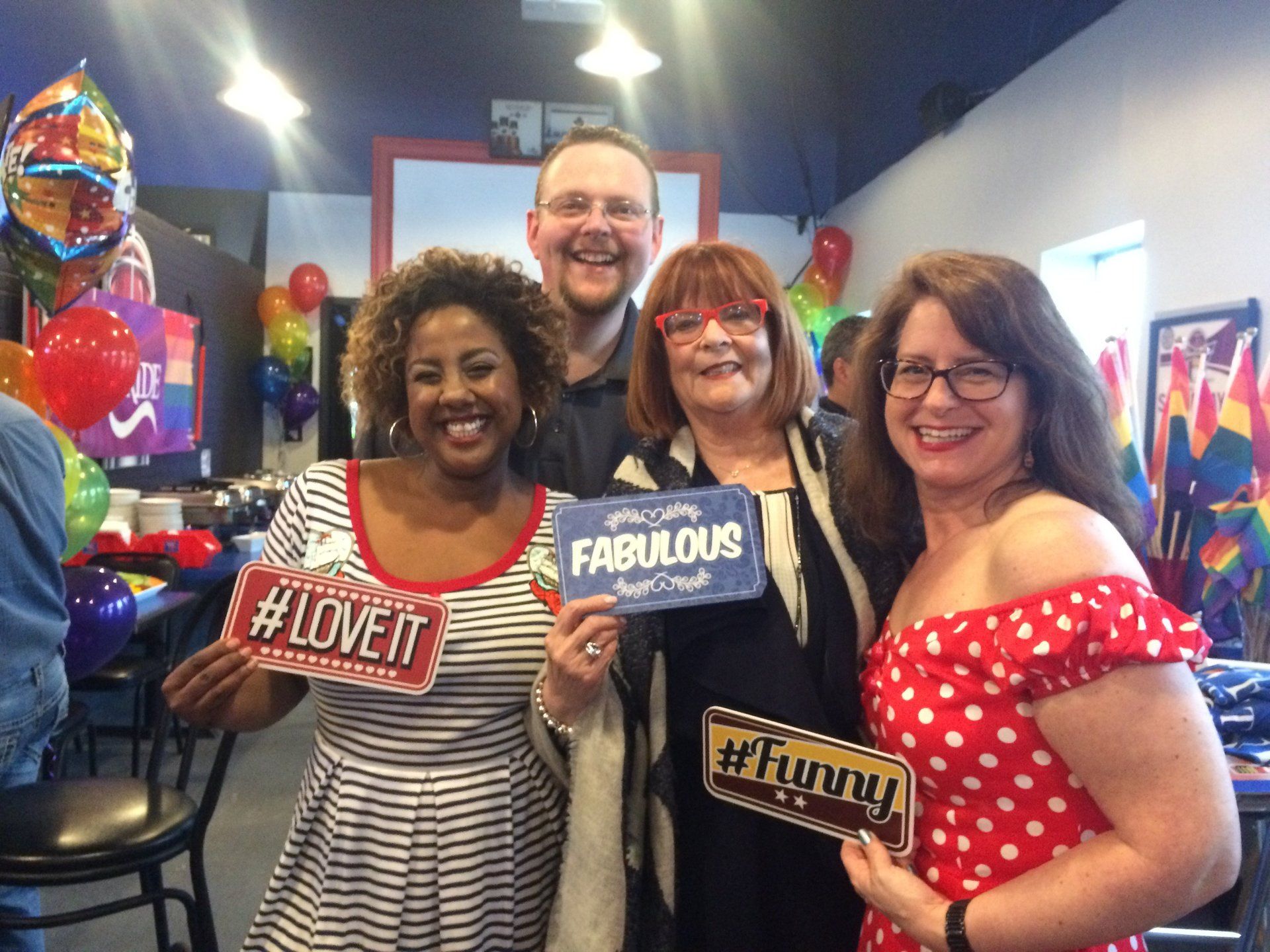 Four people pose with photo props in a colorful party setting. They smile. Balloons and art are in the background.