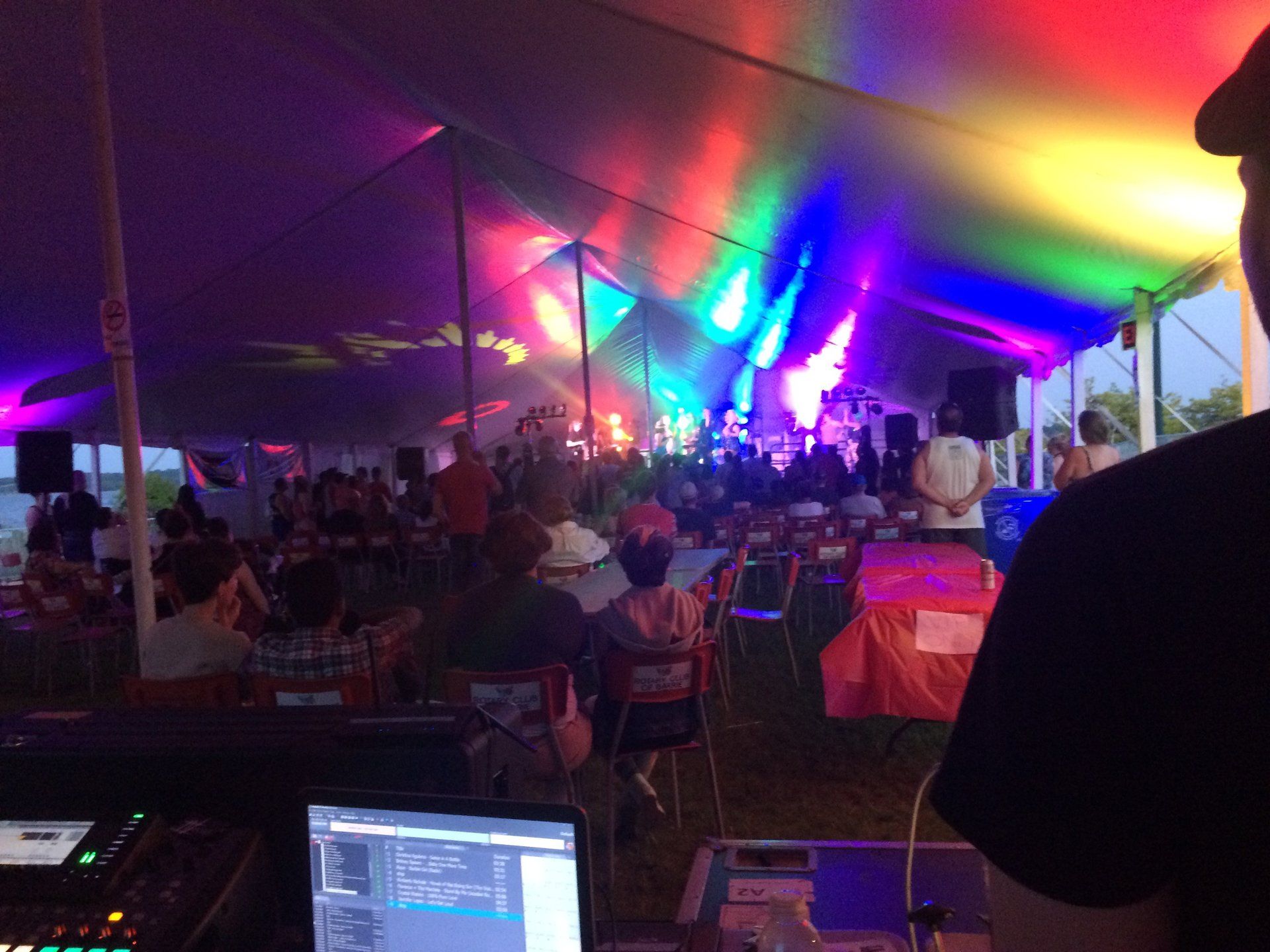 A colorful event tent interior with a band performing, people seated, and a DJ at a mixing board.