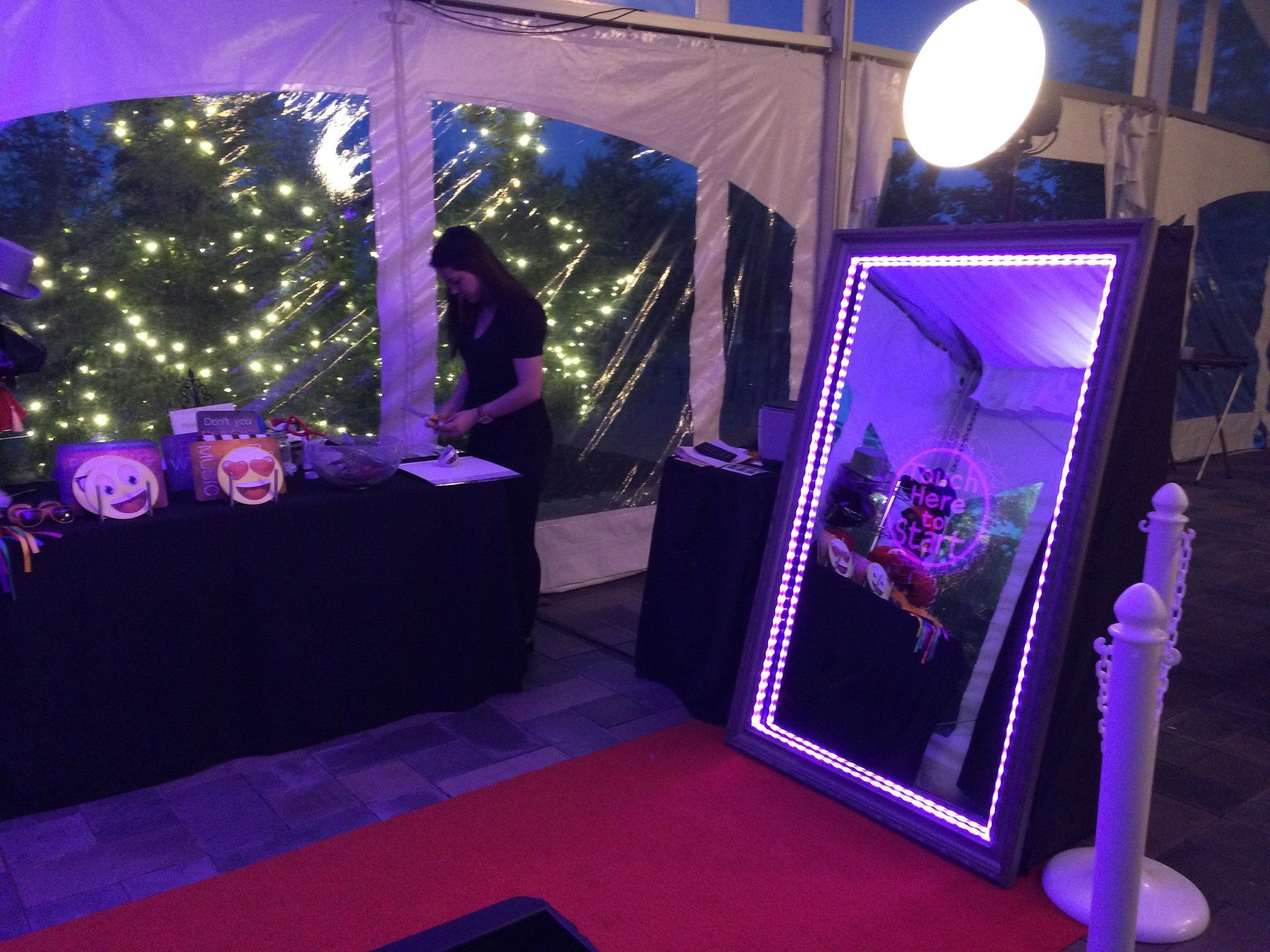Photo booth with a mirror, props, and a person setting it up under a tent with string lights.