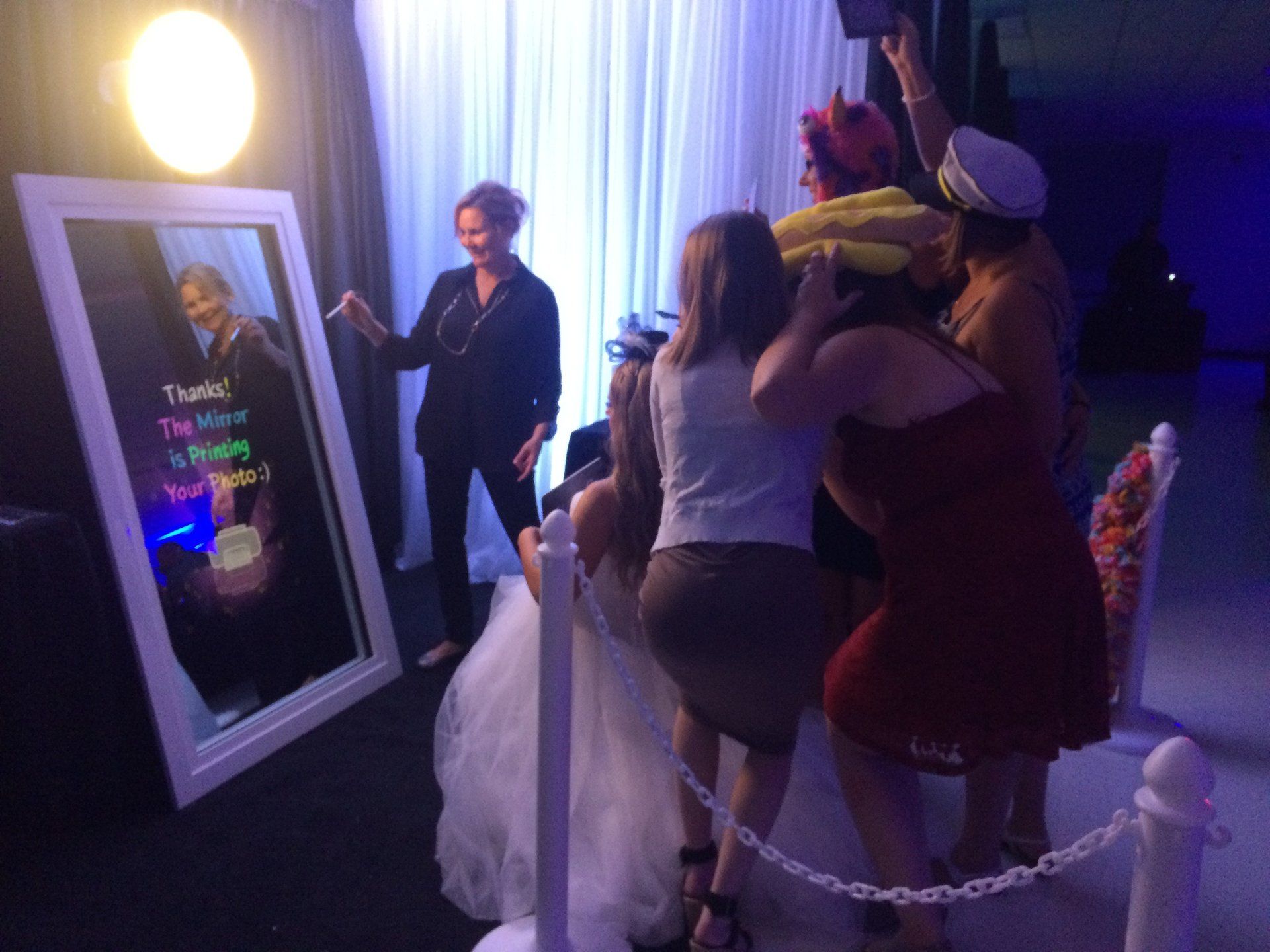 People pose with props at a photo booth mirror, a woman in a suit observes.