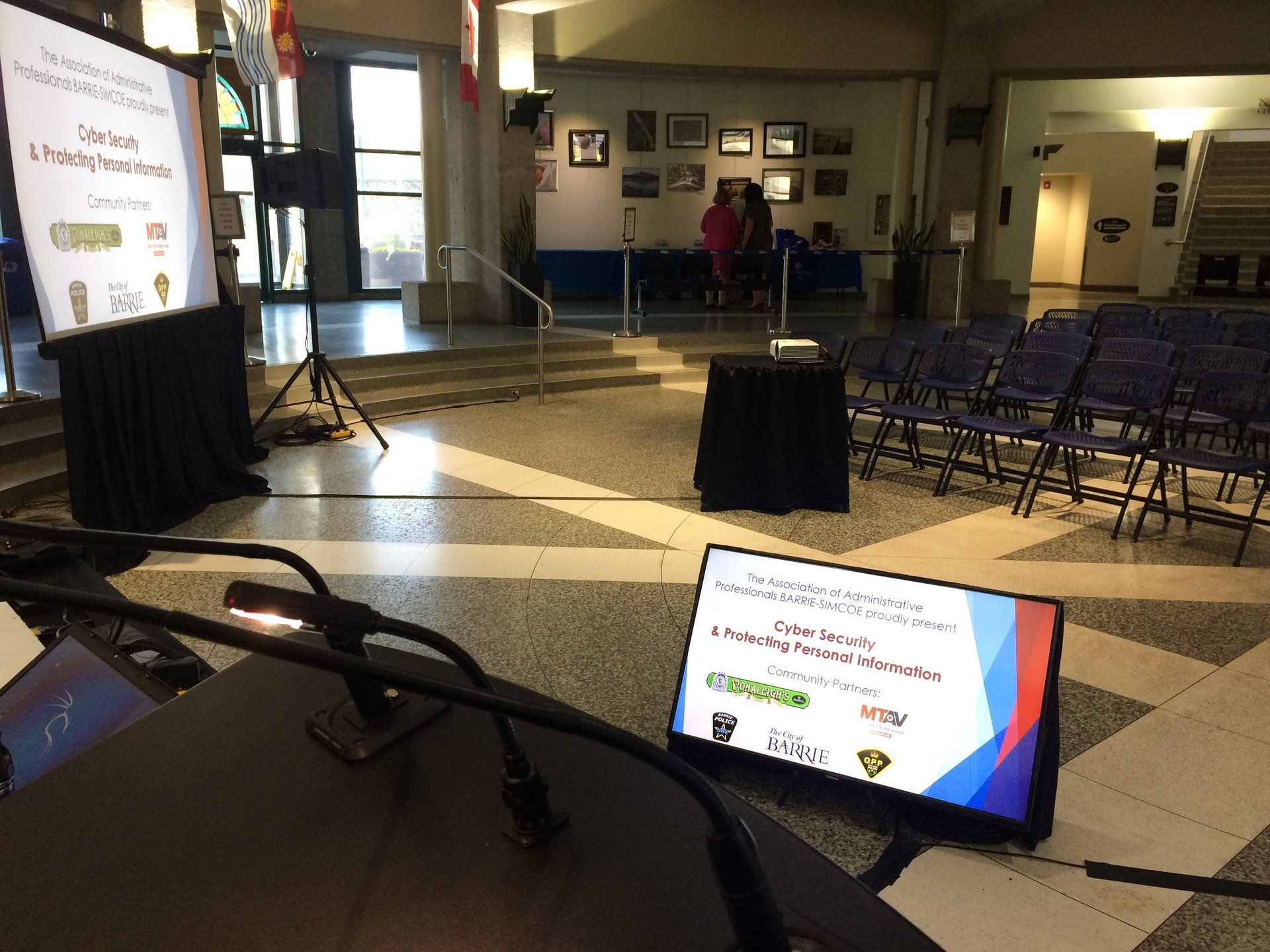 A presentation setup: microphones, screens with logos, chairs, and a person in a hall.