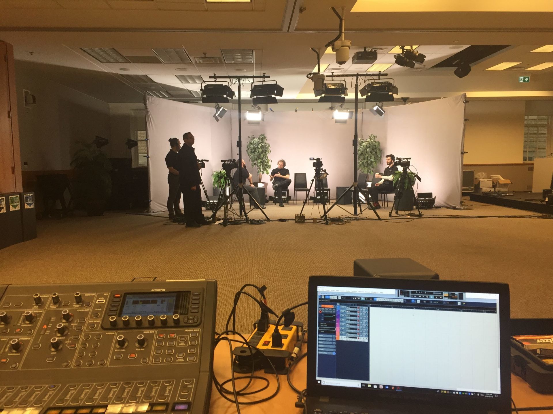 A studio setup with cameras, lights, and crew members; a soundboard and laptop in the foreground.