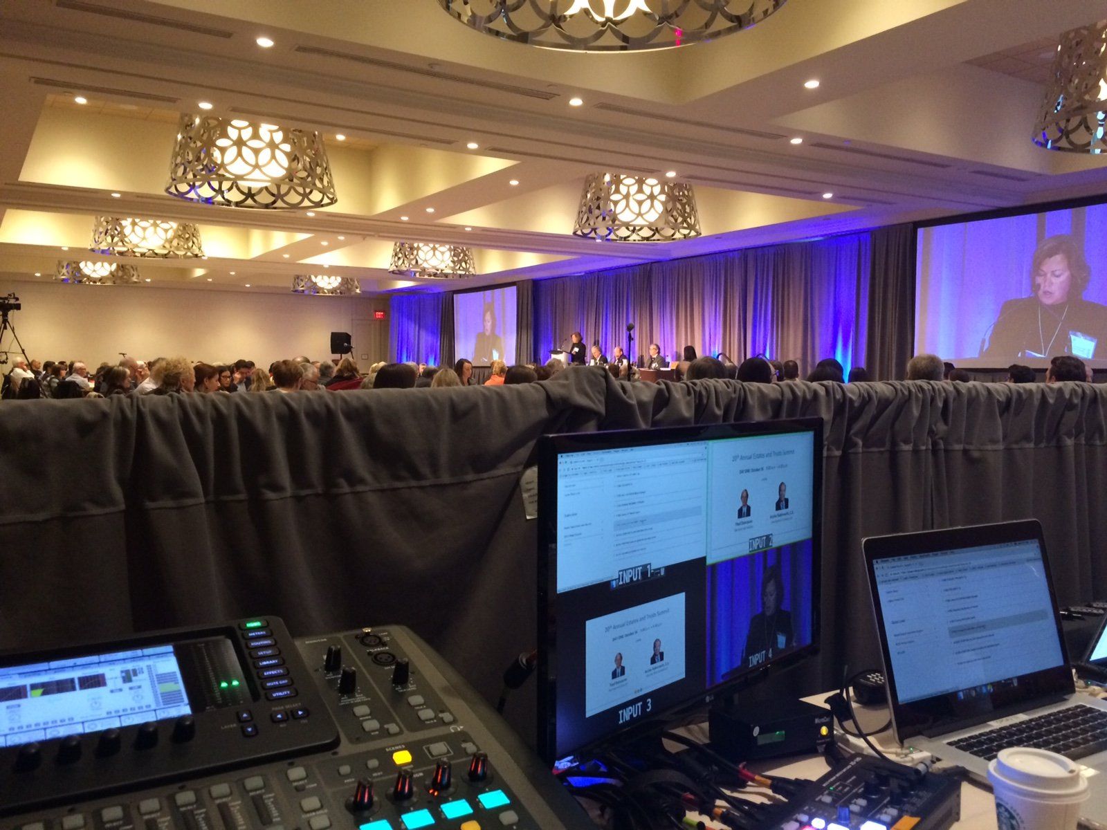 Conference with stage, screens showing presenter, and audio/video equipment in the foreground.