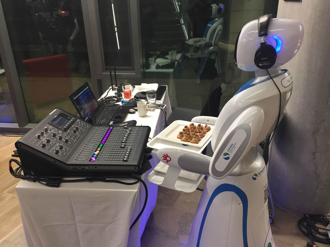 Robot DJ with headphones, holding a plate of food, standing next to a mixing board and laptop in a room.
