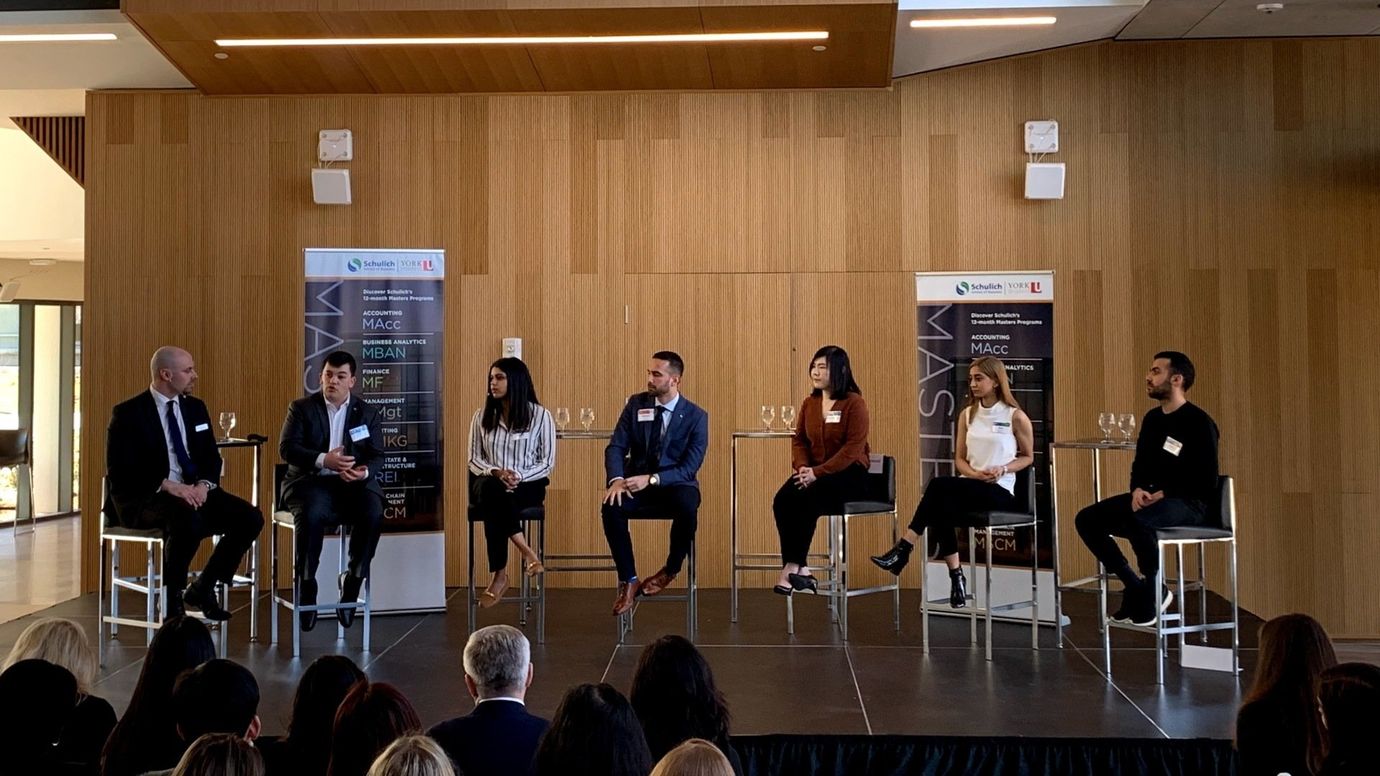 Panel discussion on stage; six people sit and speak. Audience is visible in foreground. Wooden wall backdrop.