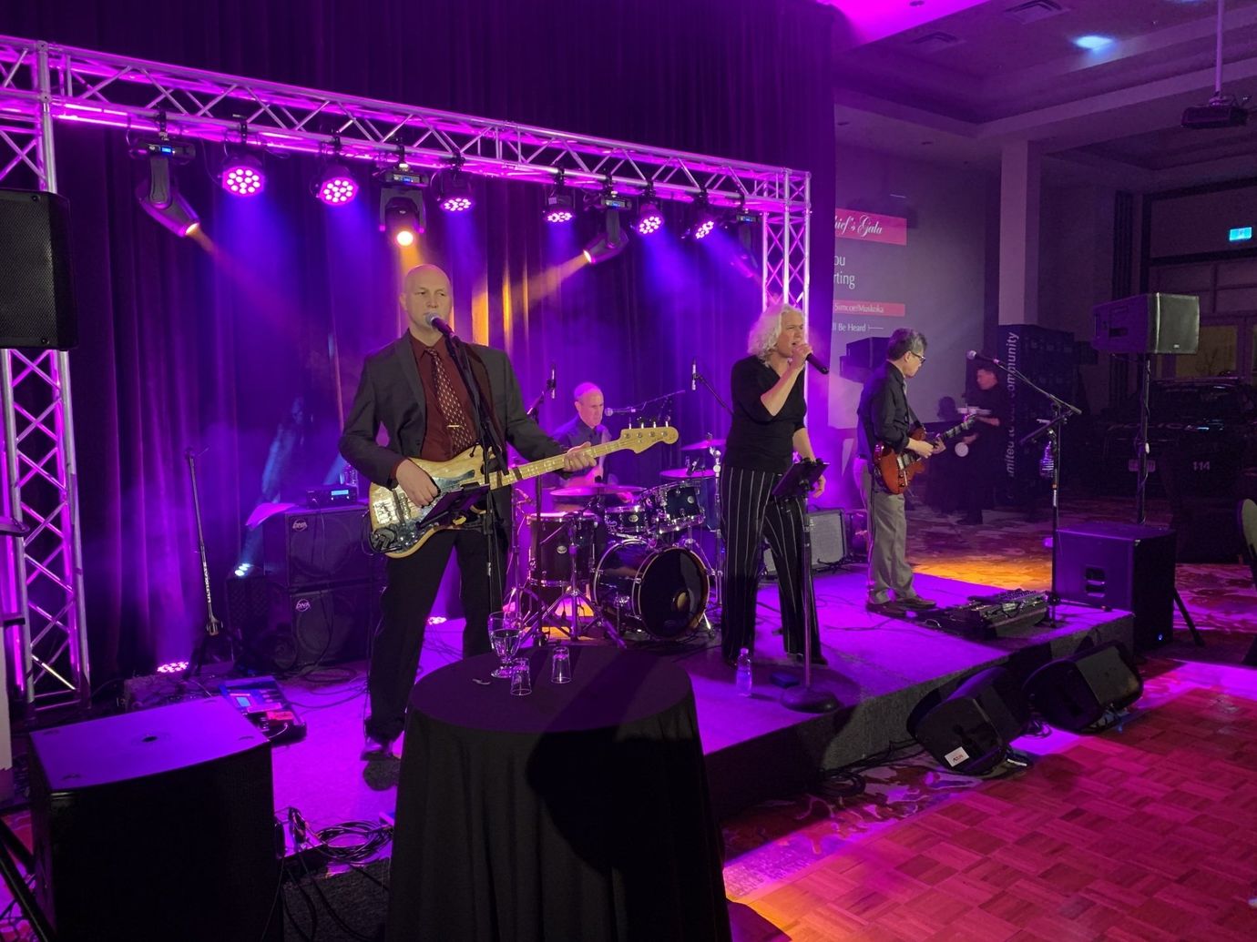 Band performing on stage under purple lights at an event. Musicians play instruments: bass, drums, guitar, and vocals.