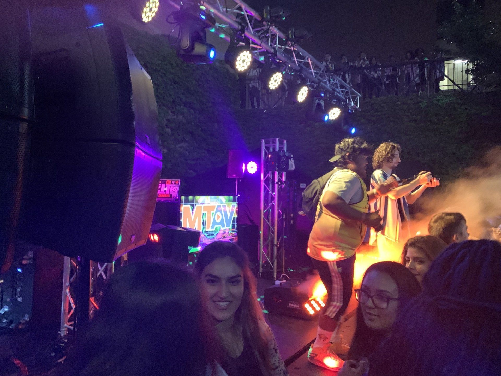People at an outdoor concert, two performers on stage with lights and crowd, night setting.