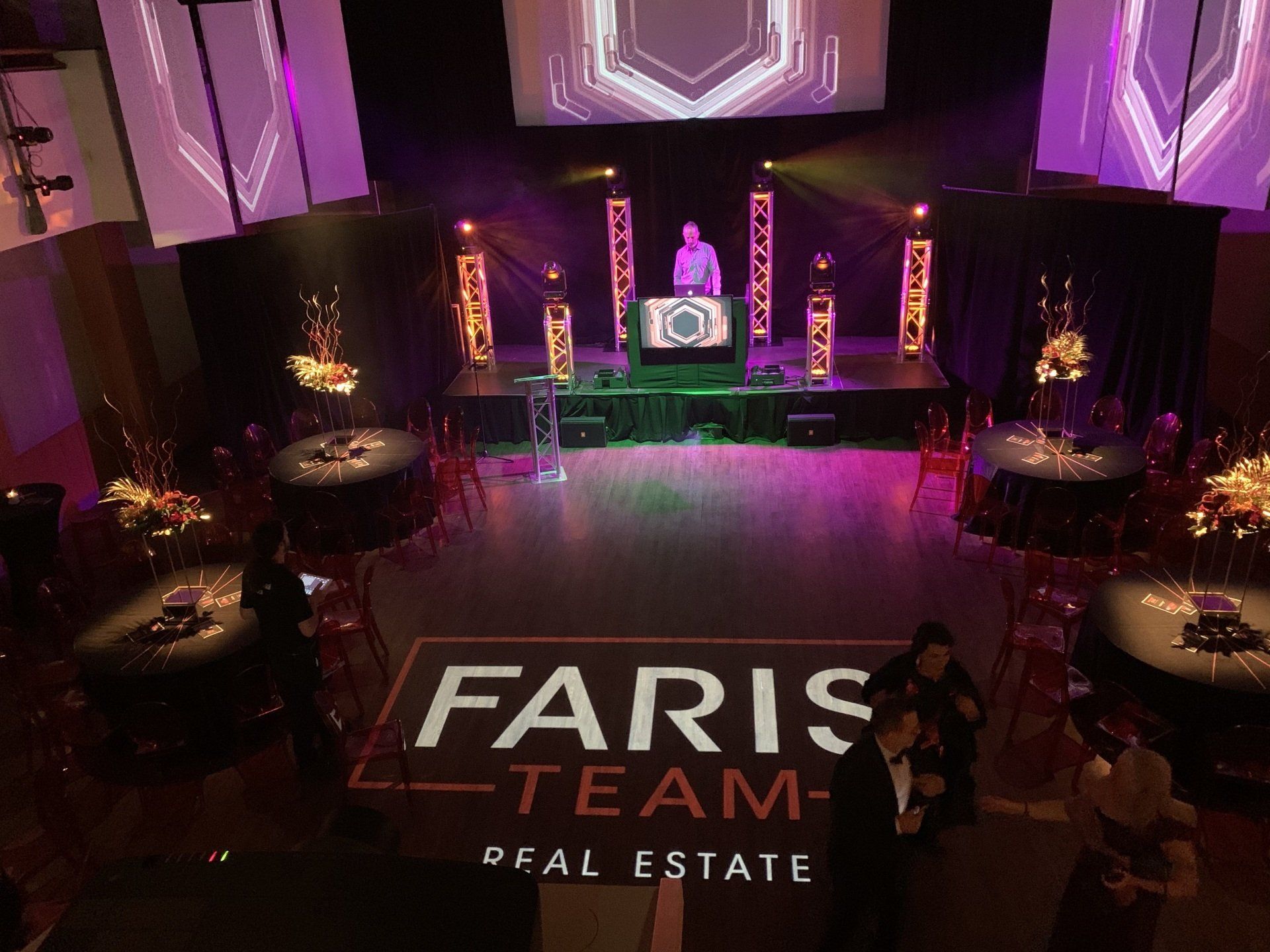 DJ on stage at a Faris Team Real Estate event with logo on dance floor, tables set for guests.