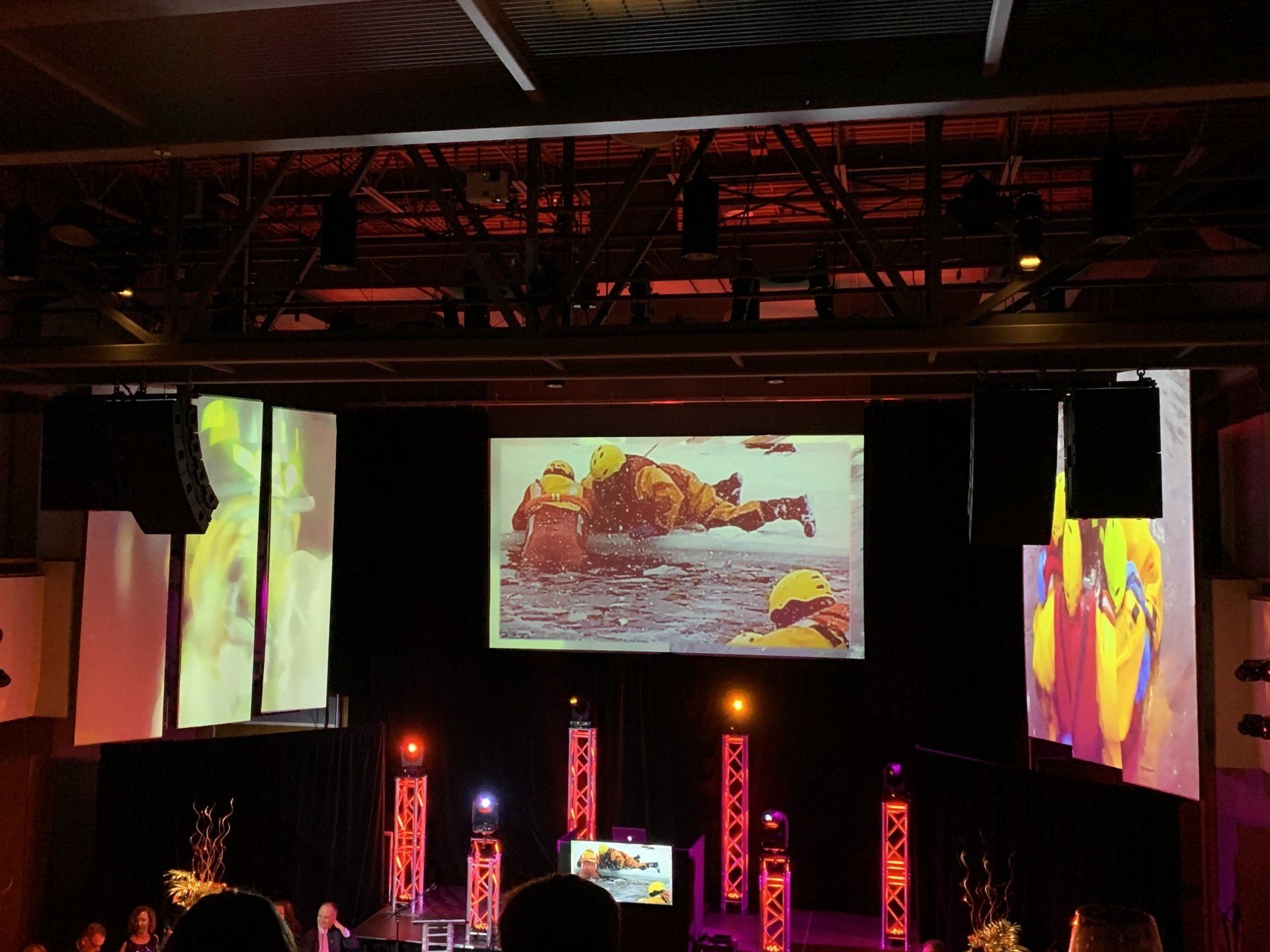Stage with central screen and side screens displaying images. Red and white lights illuminate the scene.