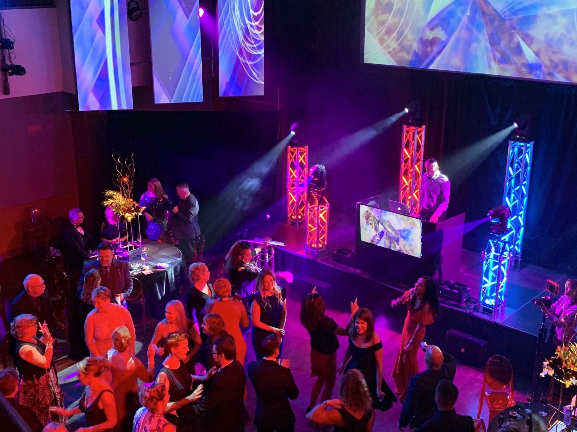 People dancing at a party with a DJ stage lit with colorful lights.