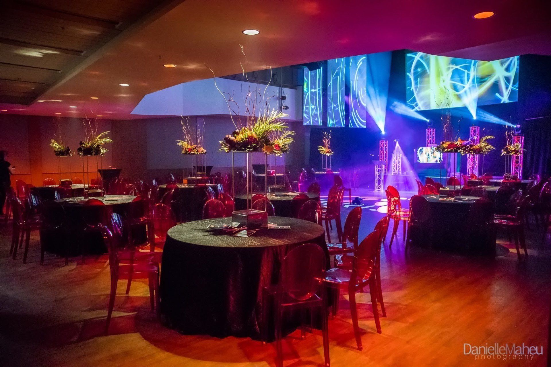 Event hall with round tables, red chairs, floral centerpieces, stage with lights.