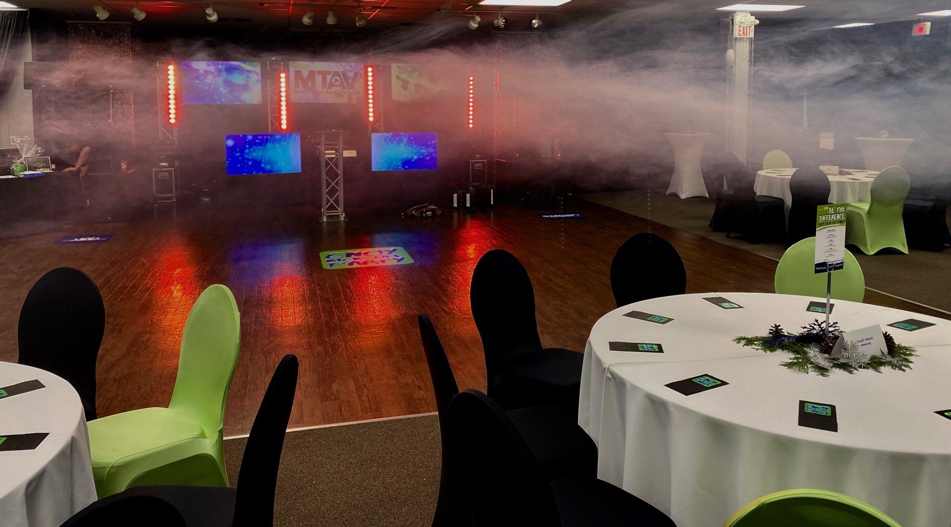 Event space with stage, tables, chairs, and smoke. Lighting is red, blue, and green.