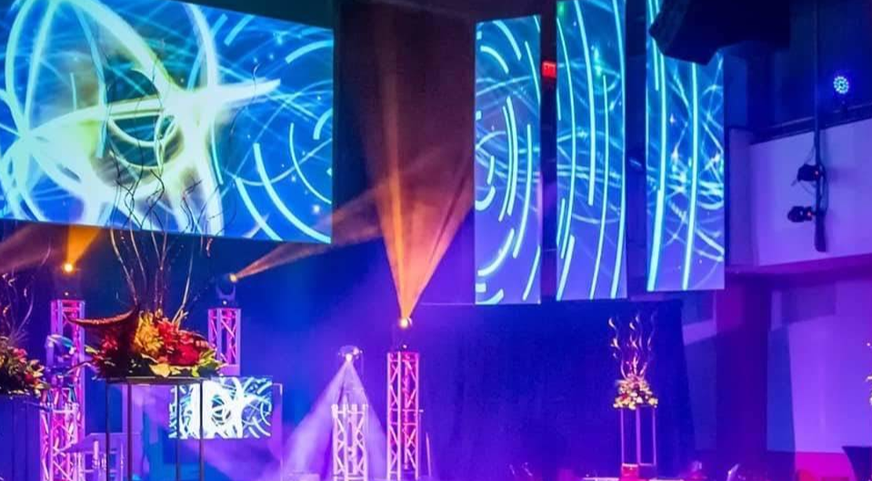 Stage with abstract blue and gold projected visuals, lights, and floral decorations.