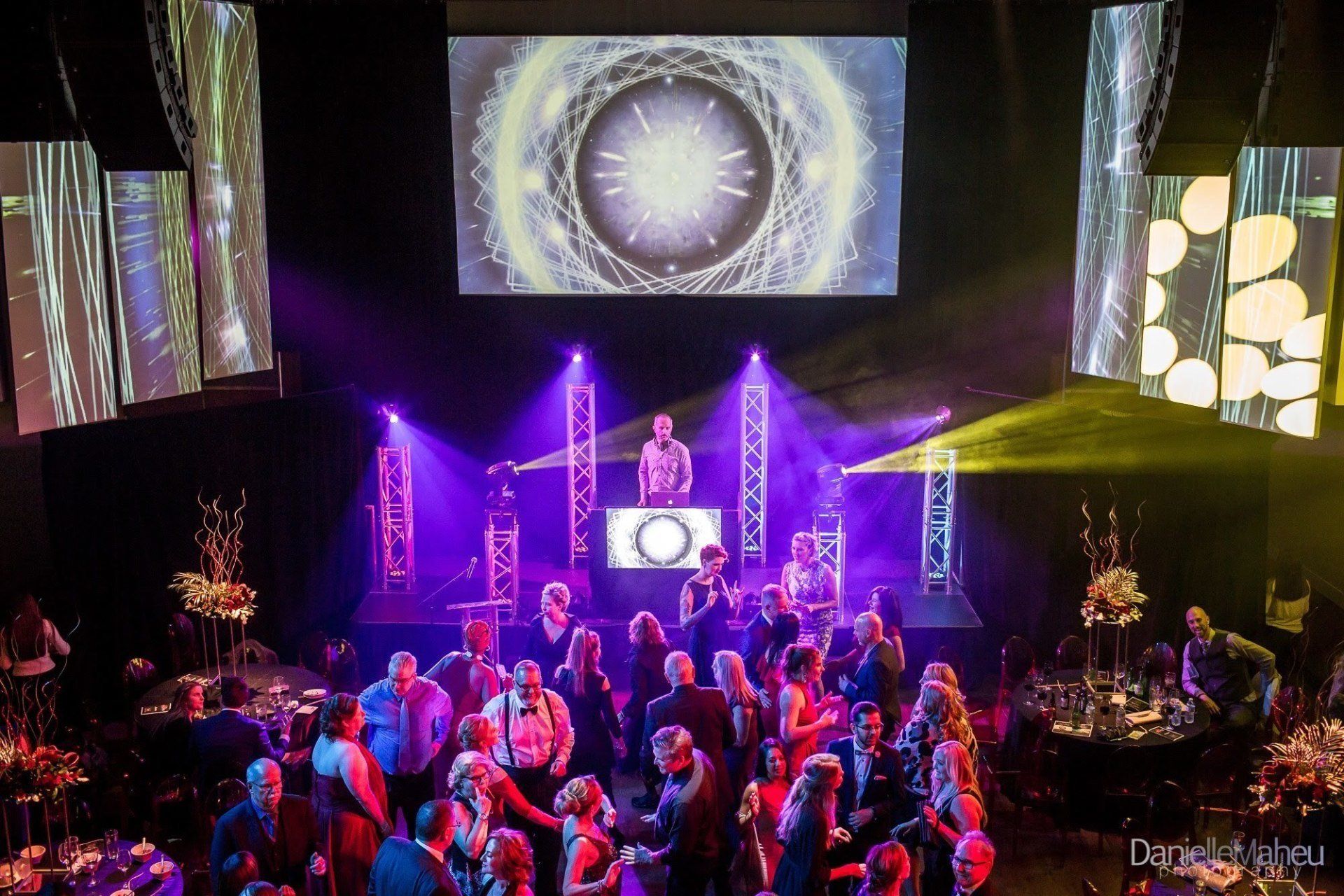DJ performing at a party with a large screen displaying abstract art, people dancing.