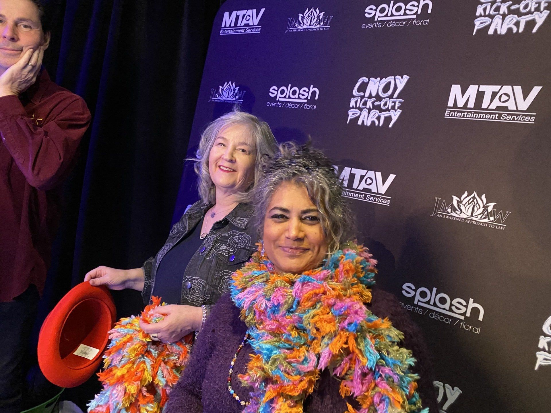 People at a party pose in front of a backdrop with logos. One woman wears a colorful scarf, another holds a red hat.