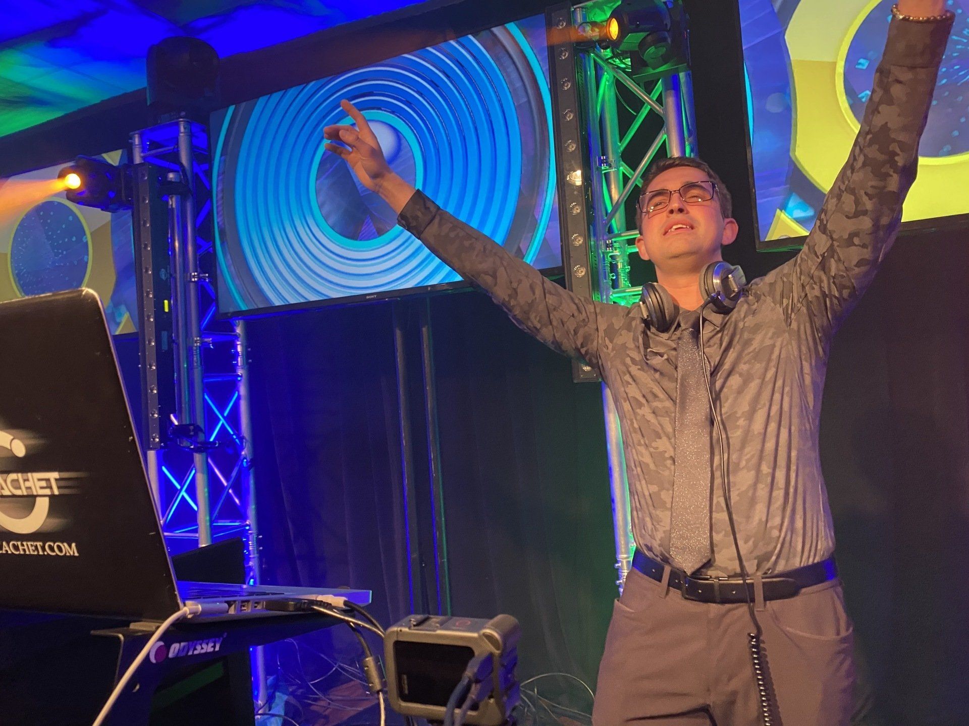 DJ at a club, arms raised. He is wearing glasses, a patterned shirt, and headphones. Blue and yellow lights behind.