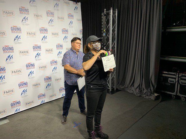 Woman in cap and mask holds certificate and item, man behind. Event backdrop.