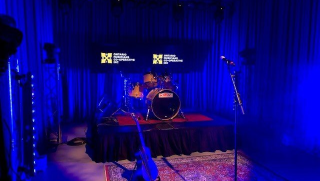 Drum set on stage under blue lights with mics, a guitar, and a logo backdrop.