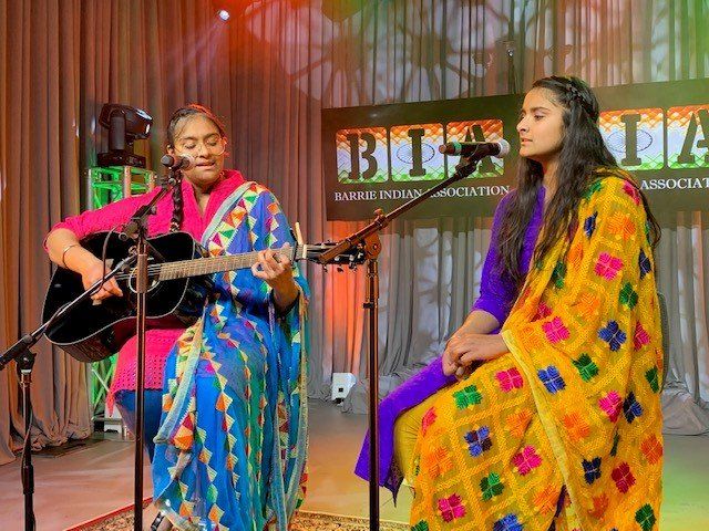 Two women singing on stage, one playing guitar. Both wear traditional colorful clothing. Stage backdrop reads 