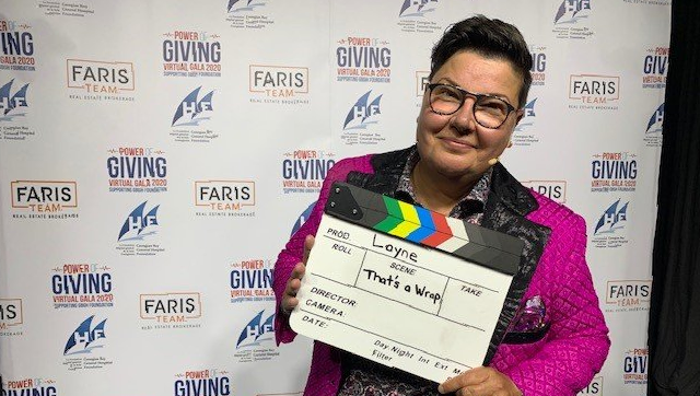 Person holding a clapperboard, smiling, in front of a banner. Wearing glasses and a pink sparkly jacket.
