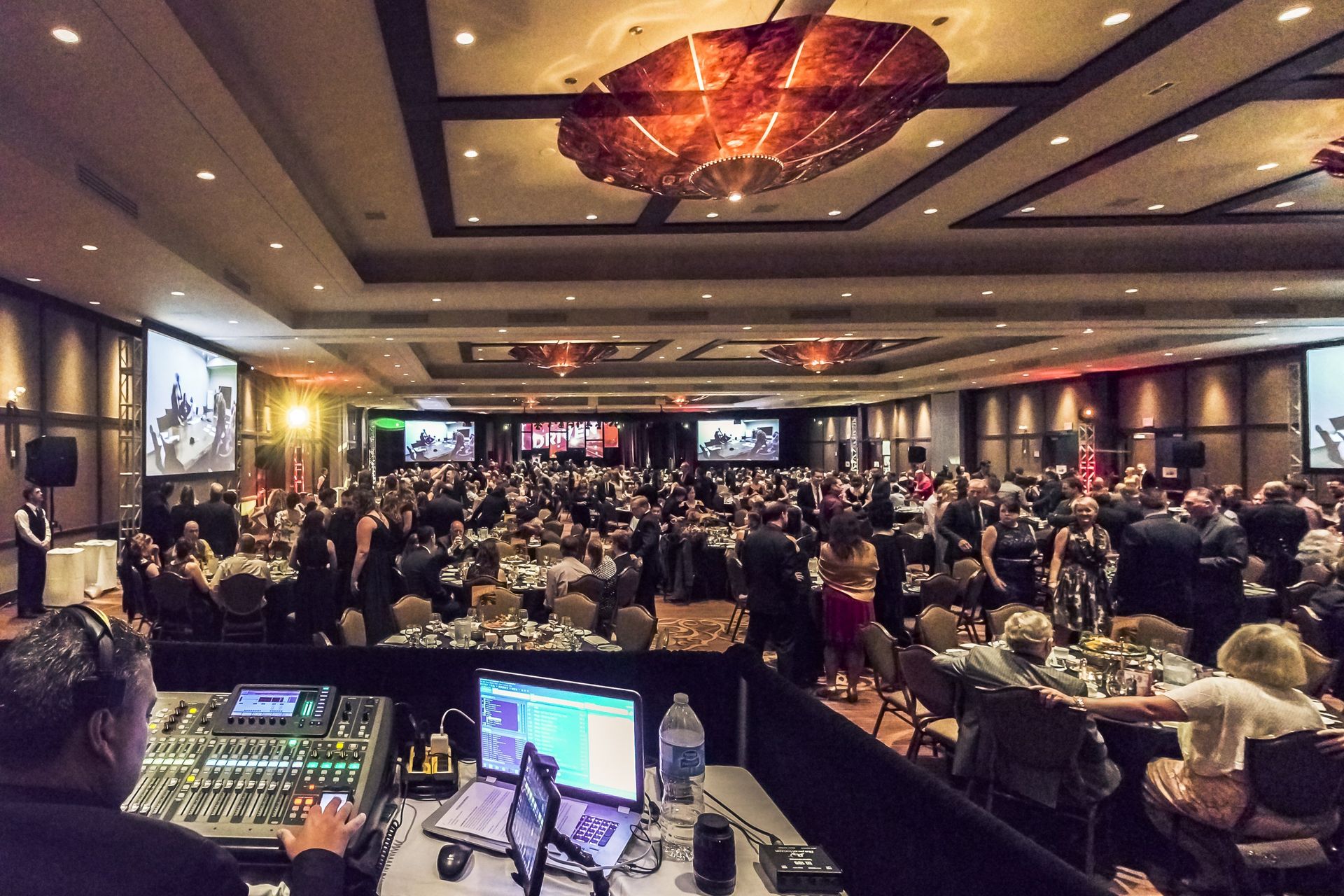 Large event in a ballroom with many people gathered. A DJ controls the soundboard in the foreground.