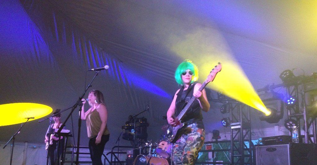 Band performing on stage with blue/yellow lighting. One guitarist with green wig, two vocalists.
