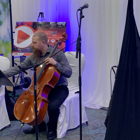 A man playing a cello, seated on a white chair, stage with microphones and music stands.