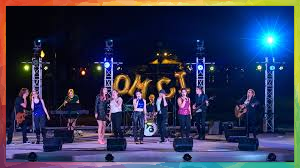 A musical ensemble performs on an outdoor stage at night, lit by stage lights and a large logo.