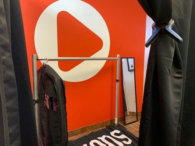 Red wall with a white play button logo, a clothing rack with a jacket, and a tall mirror. Black curtains frame the shot.