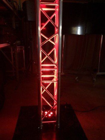 Red-lit metal truss tower on a black base. Diagonal supports create an X pattern, with light illuminating the interior.