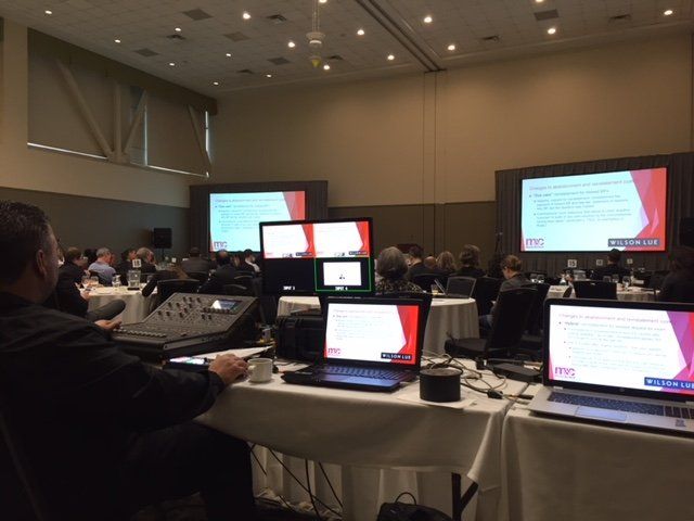 A conference room with attendees, multiple screens displaying content, and a person operating tech equipment.