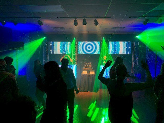 People dancing in a brightly lit room with green and blue stage lights and a DJ setup.