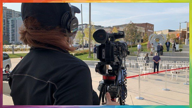 Videographer films an outdoor event. Rows of white chairs and a red ribbon are visible, along with people in the background.