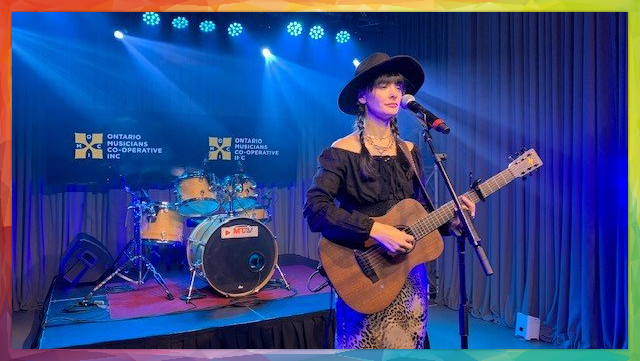 A person plays guitar and sings on stage with a drum set backdrop, blue stage lights.