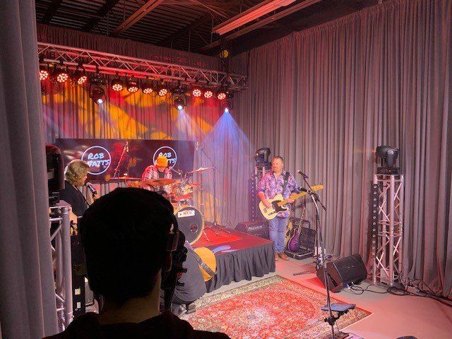 Band on stage performing under stage lights. Drums, guitar, and bass visible, with a person watching in foreground.