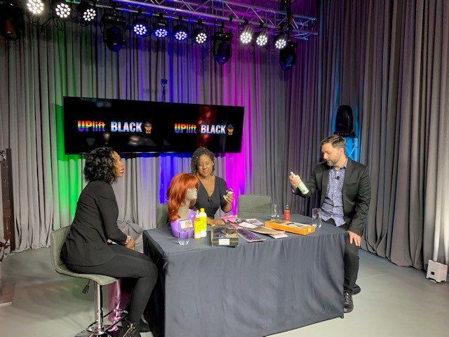 A television studio set with three people at a table.  A host gestures toward hair products. Background lights are green, purple and white.