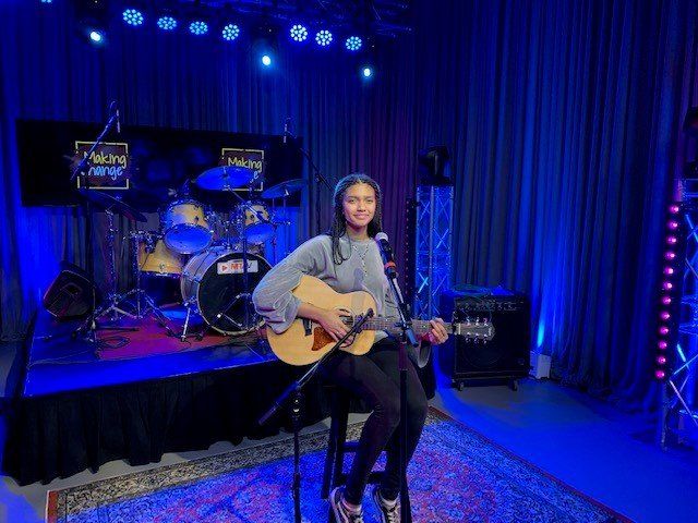 Woman playing acoustic guitar on stage in front of a drum set, microphone, and amplifier. Blue lighting.