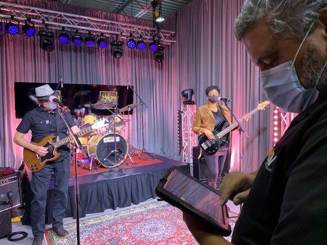 Band playing on stage, wearing masks. Crew member in foreground with tablet. Stage has lights and curtains.