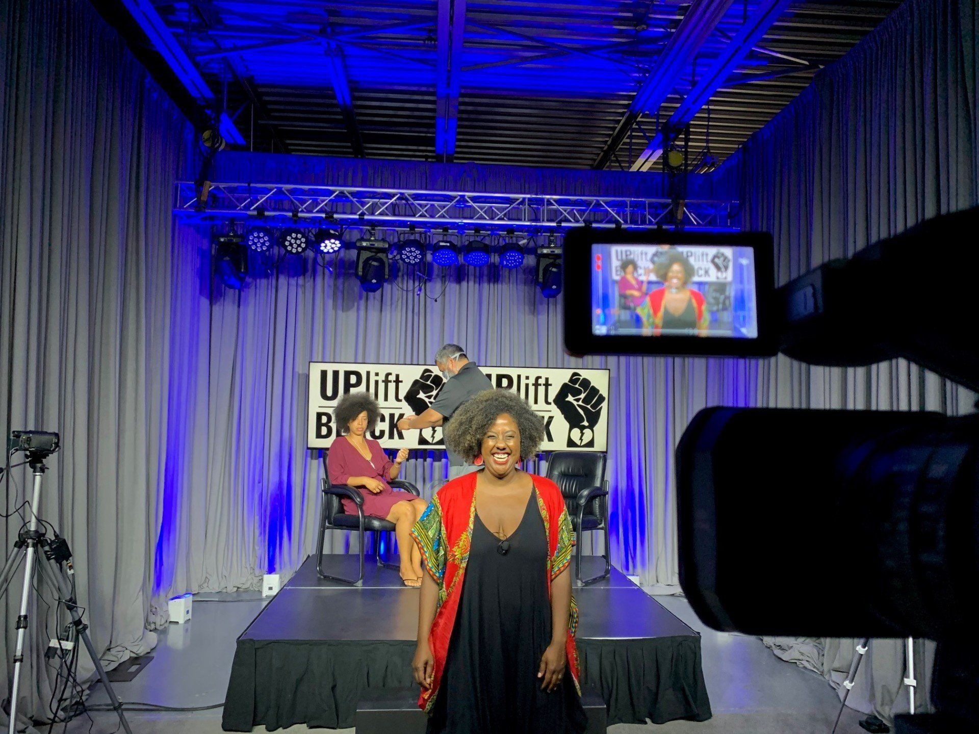 A woman in a red jacket stands on a stage; a screen shows a video with two other people; filming in a studio.