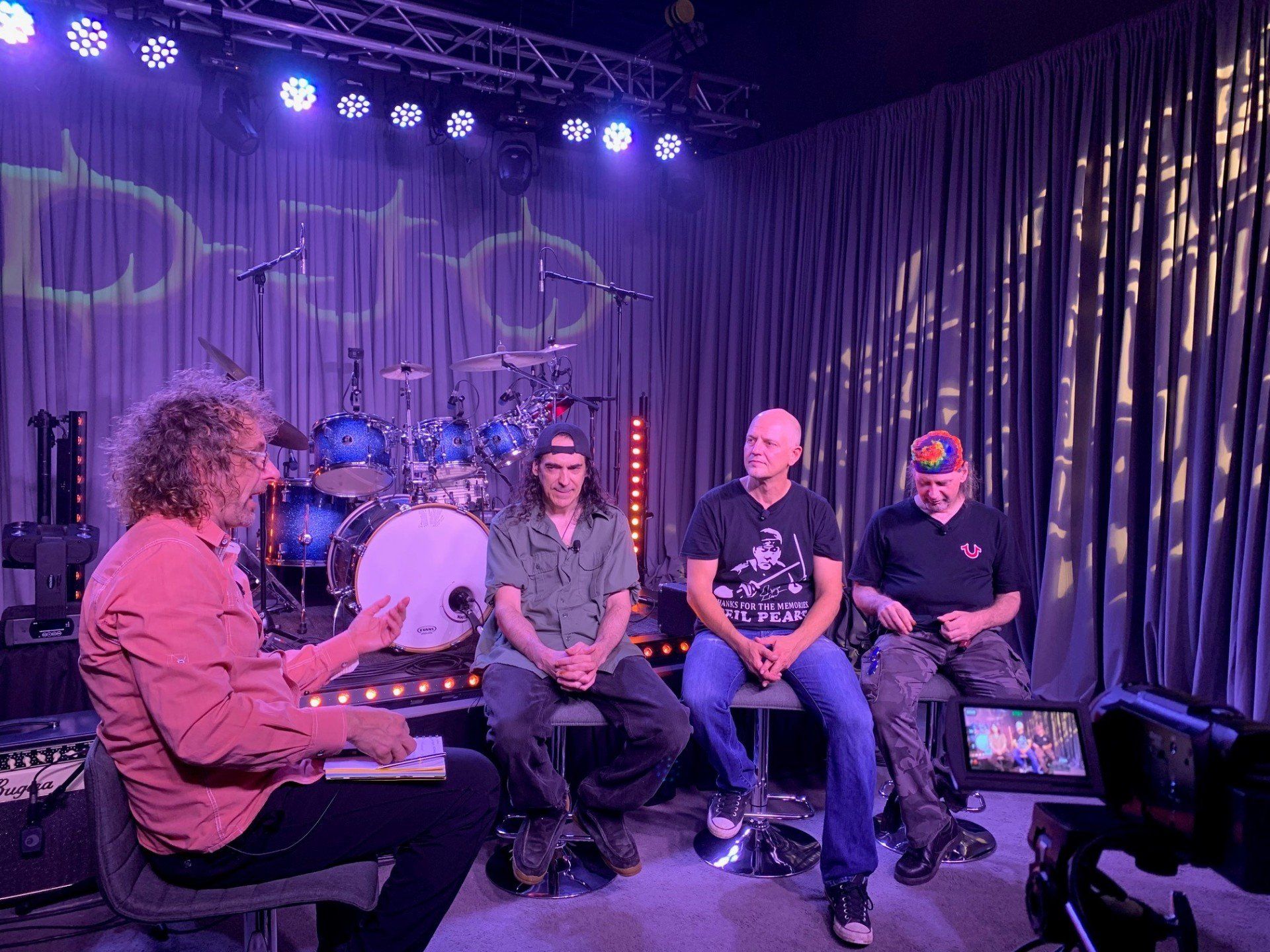 Interview with a band onstage. Four people seated, a drummer behind them, lit by purple stage lights.
