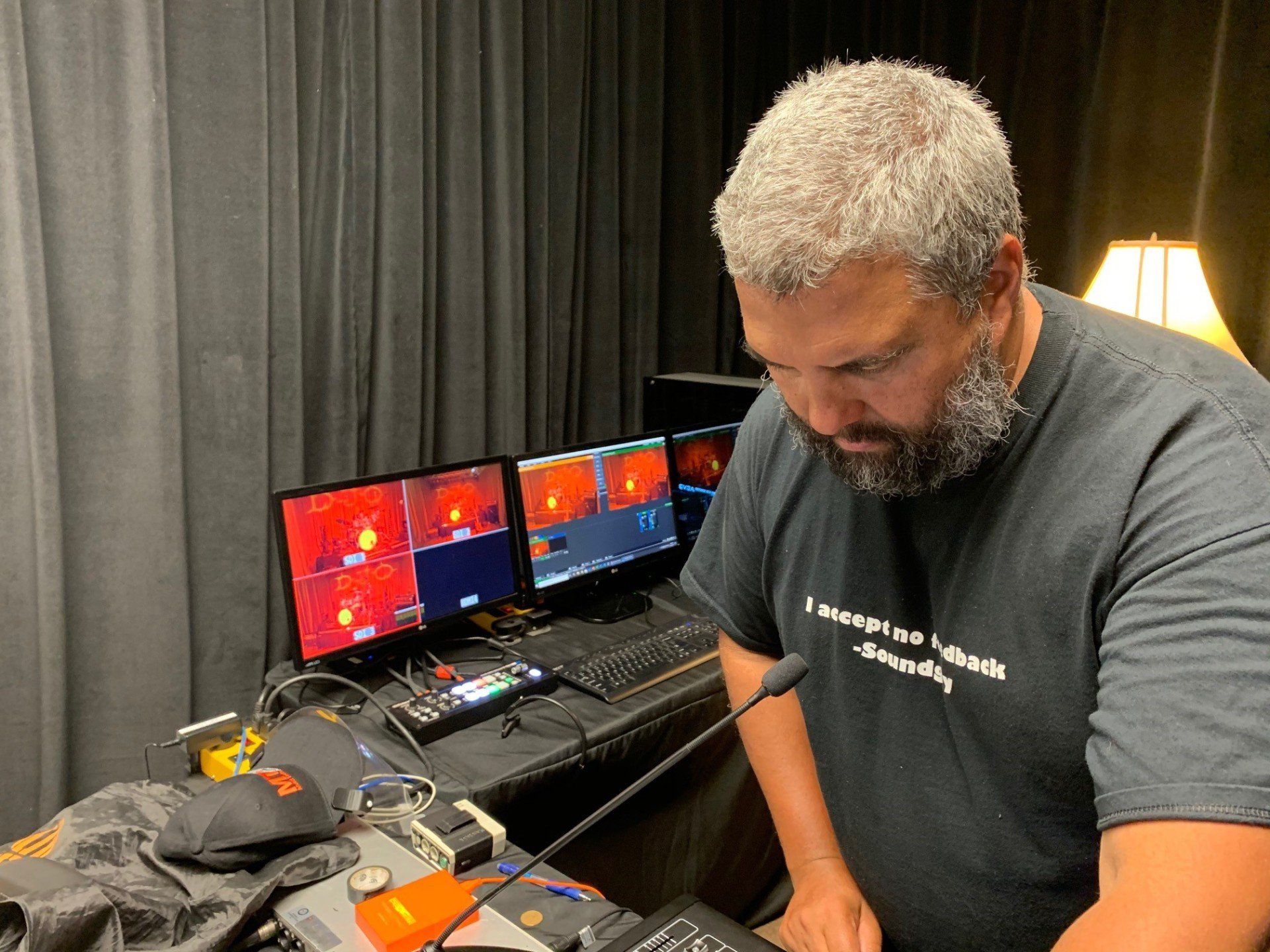 Man in black t-shirt looking at monitors with red screens and equipment. A microphone is in front of him.