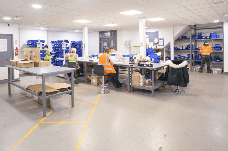 Warehouse workspace with workers assembling items at tables, blue bins stacked along the walls.