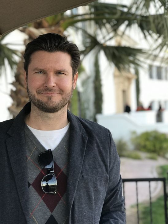 Jesse Clark Bio Pic a man with a beard and sunglasses is standing in front of a palm tree. His name is Jesse Clark