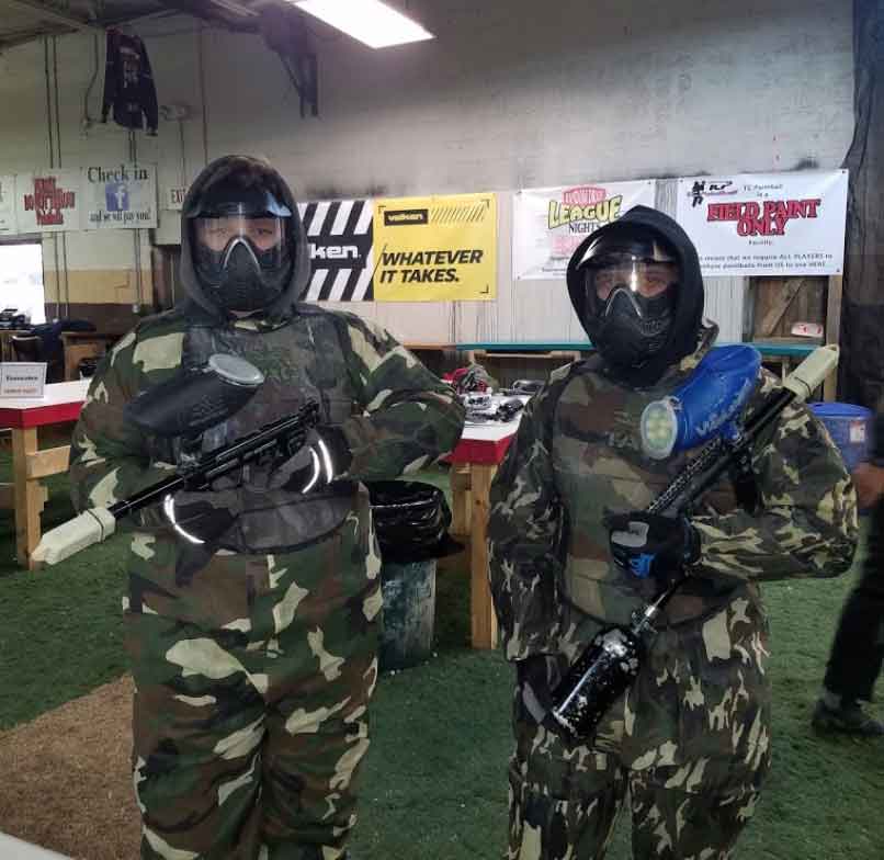 Two paintball players stand in front of a sign that says whatever it takes