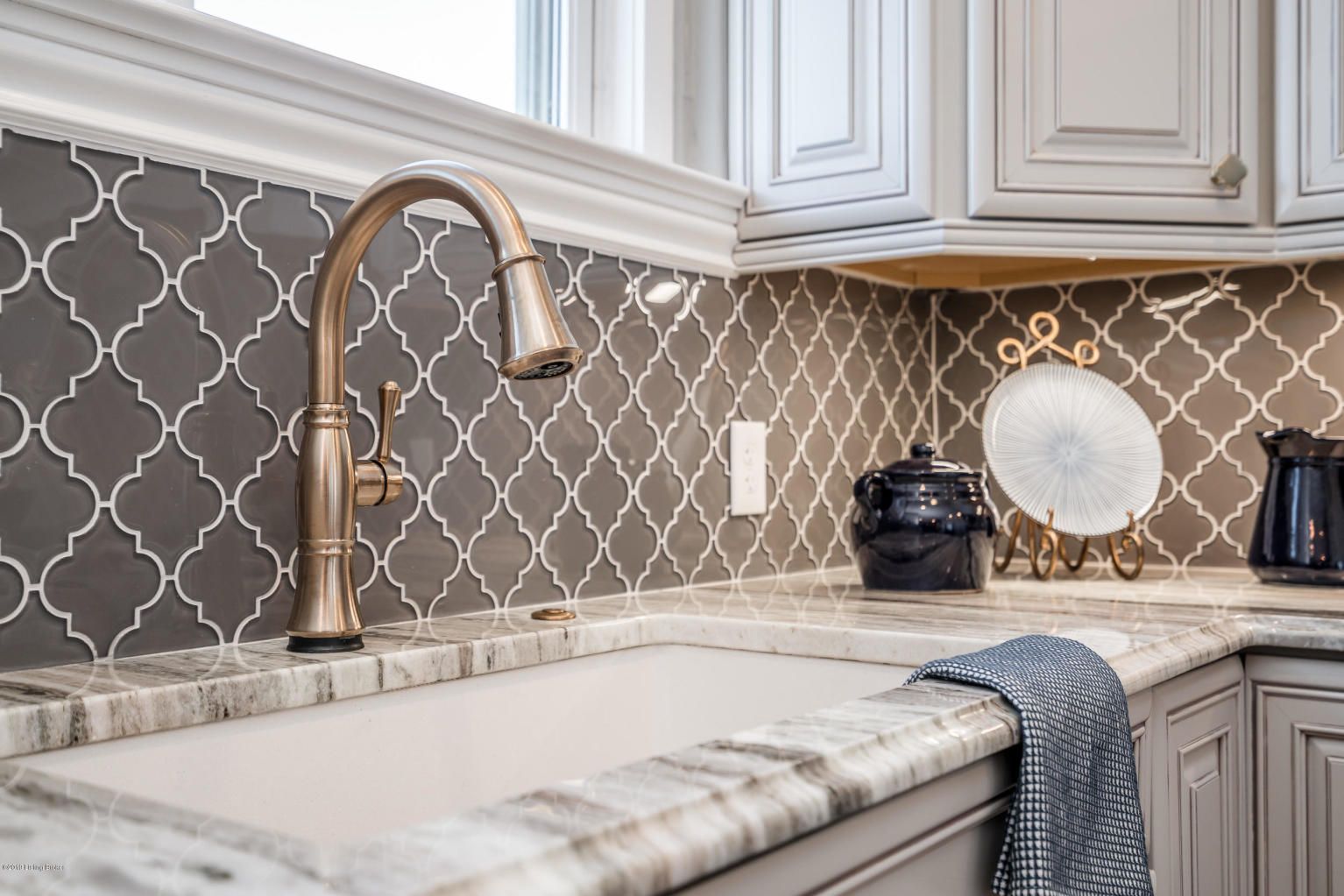 Close-up of a kitchen sink with ornate gray and white tile backsplash and bronze faucet.