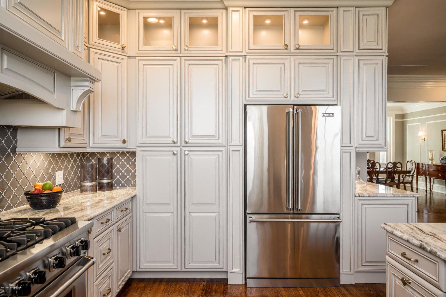White kitchen cabinets surround stainless steel refrigerator. Granite countertops and dark wood floor.