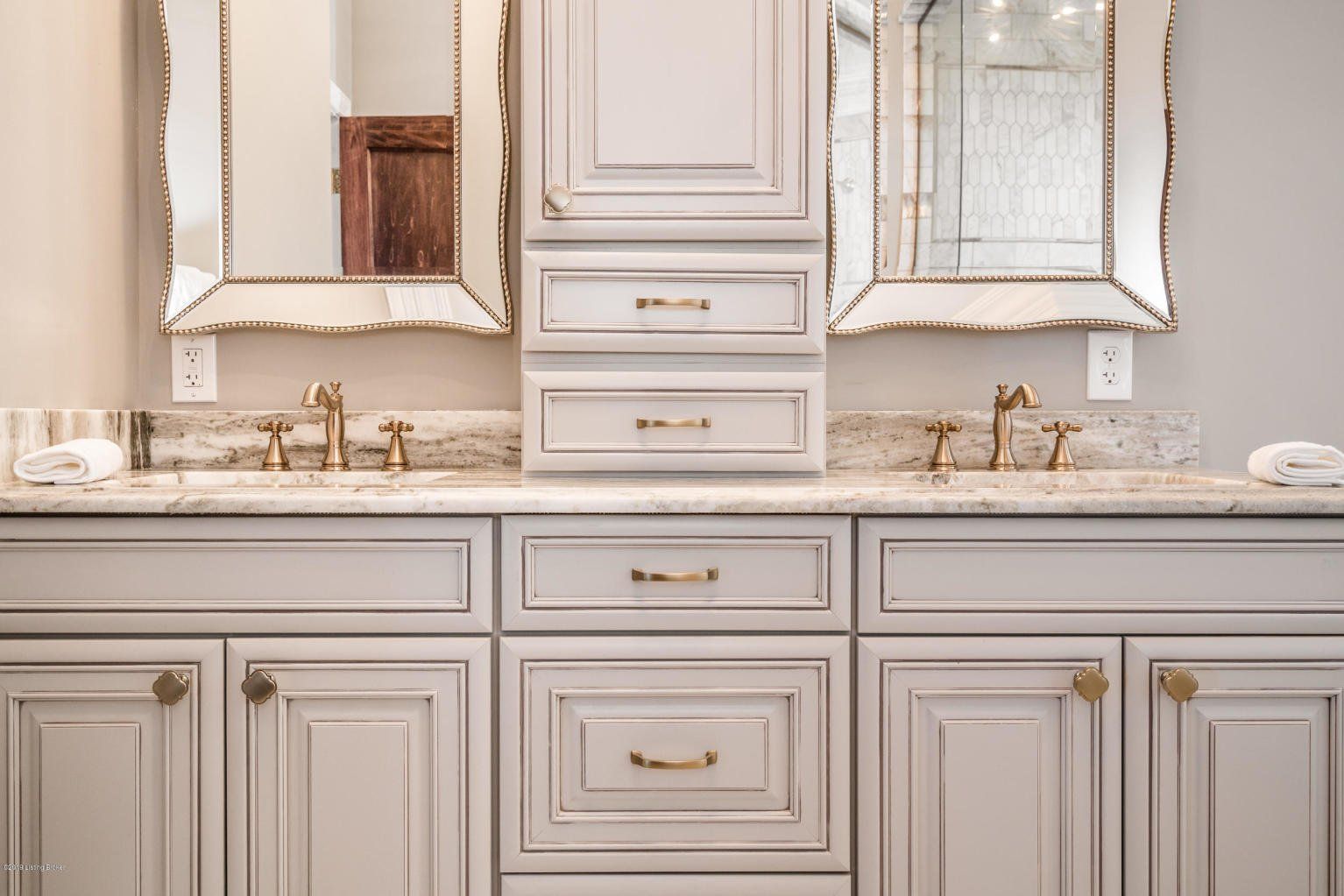 Bathroom vanity with two mirrors, sinks, and a central storage cabinet; neutral color scheme with gold accents.