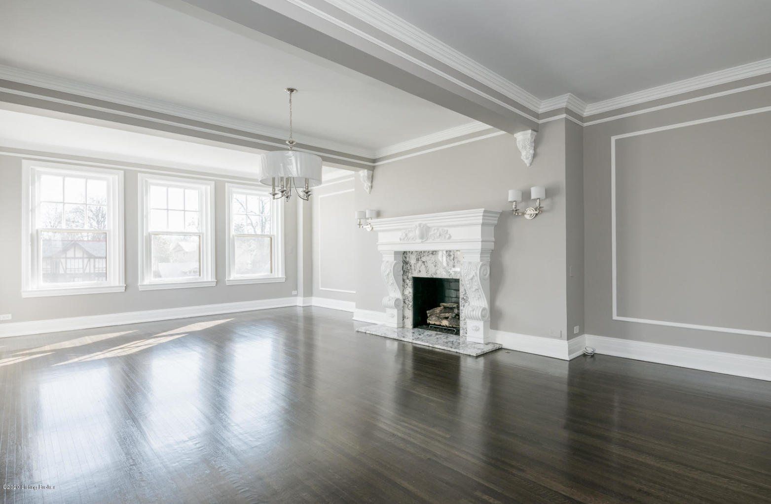 Empty, light-filled living room with dark hardwood floors, white fireplace, and chandelier.